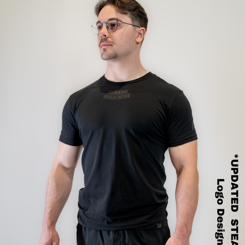 Man wearing A7's IPF Approved Stealth black Meet shirt. 'Demand Greatness’ written on the front in a dark grey double outlined font to have impact on the competition platform. Paired with black shorts made from a stretchy squat proof material to use in powerlifting training.