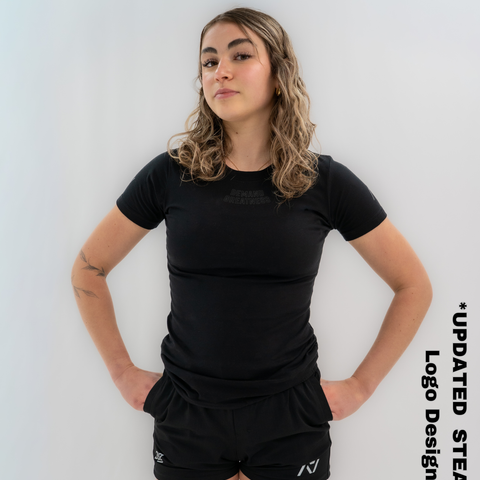Woman wearing A7's IPF Approved Stealth Meet shirt in black. 'Demand Greatness’ written on the front in a dark grey double outlined font to have impact on the competition platform. Paired with black shorts made from a stretchy squat proof material to use in powerlifting training.