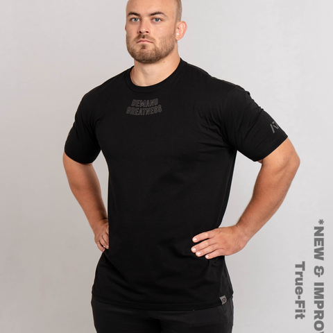 Man wearing A7's IPF Approved Shadow Stone Black Meet shirt in black. 'Demand Greatness’ written on the front in a grey double outlined font to have impact on the competition platform. Paired with black powerlifting training joggers. 