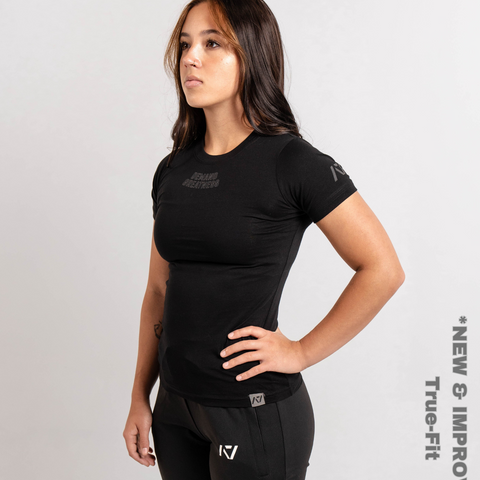 Woman wearing A7's IPF Approved Shadow Stone Black Meet shirt. 'Demand Greatness’ written on the front in a grey double outlined font to have impact on the competition platform. Paired with black powerlifting training joggers. 

