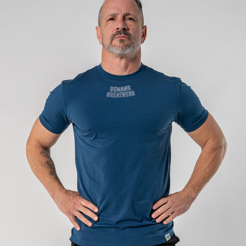 Front view of Man wearing A7's IPF Approved Tides Meet shirt in a dark blue. 'Demand Greatness’ written on the front in a white double outlined font to have impact on the competition platform. Paired with black shorts made from a stretchy squat proof material to use in powerlifting training.
