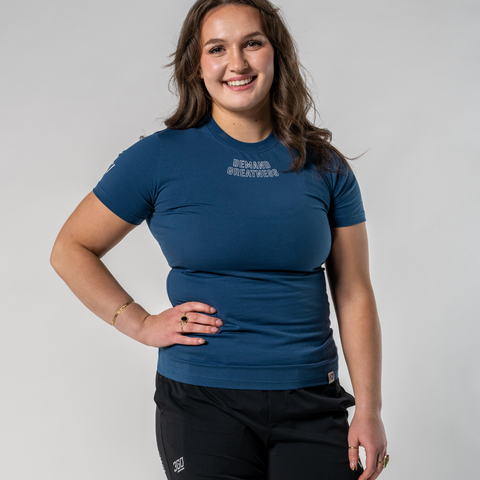 Front view of Woman wearing A7's IPF Approved Tides Meet shirt in a dark blue. 'Demand Greatness’ written on the front in a white double outlined font to have impact on the competition platform. Paired with black shorts made from a stretchy squat proof material to use in powerlifting training.