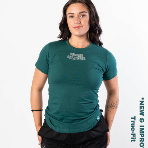 Woman wearing A7's IPF Approved Emerald Foras green Meet shirt. 'Demand Greatness’ written on the front of green meet t-shirt in a white double outlined font to have impact on the competition platform. Paired with black powerlifting shorts made from a stretchy squat proof material to use in strength sports training. 
