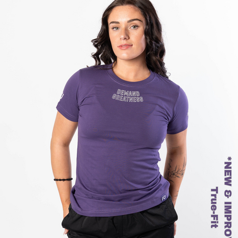 Woman wearing A7's IPF Approved Purple Meet shirt. 'Demand Greatness’ written on the front in a white double outlined font to have impact on the competition platform. Paired with black powerlifting shorts made from a stretchy squat proof material to use in strength sports training. 