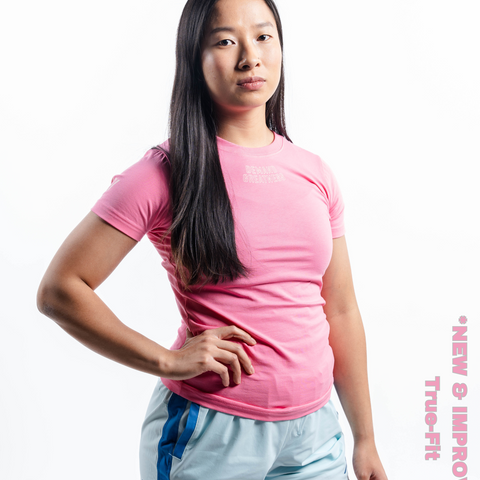 Woman wearing A7's IPF Approved Roseite light pink Meet shirt. 'Demand Greatness’ written on the front in a white double outlined font to have impact on the competition platform. Paired with light blue powerlifting shorts made from a stretchy squat proof material to use in strength sports training. 
